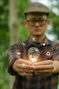 Man holding glowing light bulb with clean energy icons compared to unlit bulb in green nature background.