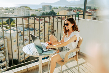 Obraz premium Smiling female freelancer in sunglasses using computer on sunlit terrace during summer vacation.