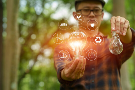 Man holding glowing light bulb with clean energy icons compared to unlit bulb in green nature background.