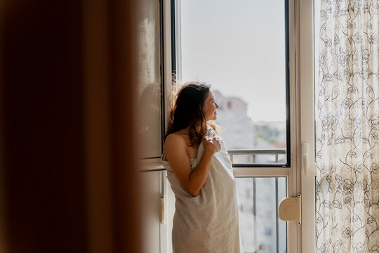 Beautiful young woman standing by window wrapped in white sheet, greeting a new day and looking at the city.