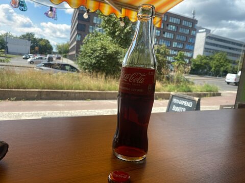 Glasflasche Coca-Cola auf einem Restauranttisch vor urbaner Stadtkulisse in Deutschland. Symbolbild f&uuml;r Softdrinks, Gastronomie, Markenprodukte und Alltag. Nur f&uuml;r redaktionelle Nutzung.