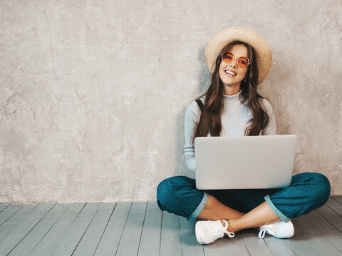 Portrait of creative young smiling woman in sunglasses. Beautiful sitting on the floor near gray wall. Model using notebook. Female dressed in hipster clothes and hat