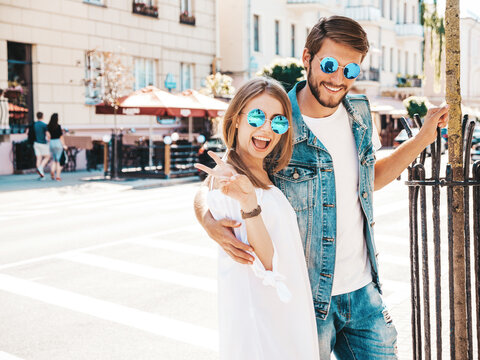 Smiling beautiful female and her handsome boyfriend posing in the street. Woman in casual summer dress and man in jeans clothes. Happy cheerful couple family having fun in sunglasses.Shows peace sign