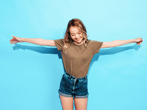 Young beautiful woman looking at camera. Trendy in casual summer shirt clothes with natural makeup. Positive female smiling. Funny model posing near blue wall in studio.Raises her hands