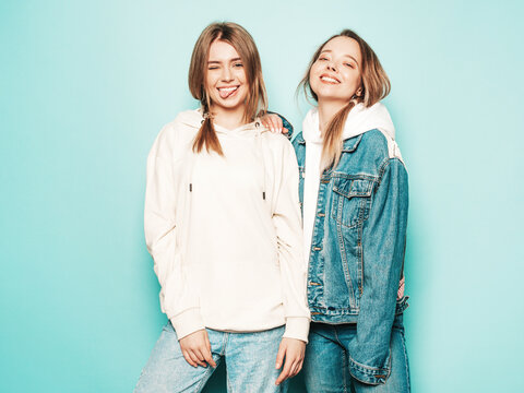 Two young beautiful brunette smiling hipster in trendy summer hoodie and jeans jacket clothes. Sexy carefree women posing near blue wall. Trendy and positive models having fun
