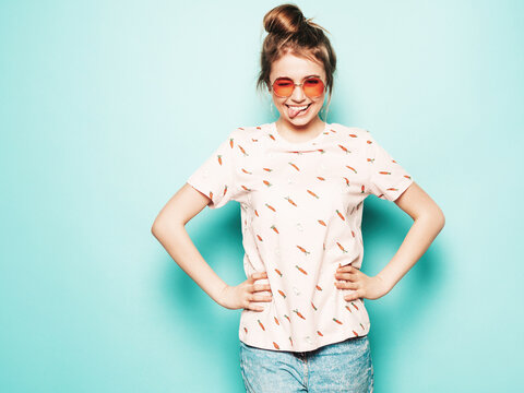 Young beautiful woman looking at camera. Trendy in casual summer T-shirt clothes showing her tongue. Positive female shows facial emotions. Funny model isolated on blue in sunglasses