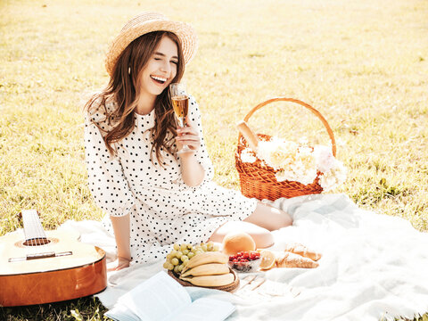 Young beautiful hipster woman in trendy summer sundress and hat. Carefree woman making picnic outside. Positive model sitting on plaid on grass, eating fruits and cheese, drinking champagne