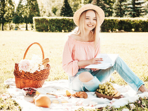 Young beautiful hipster woman in trendy summer jeans, pink T-shirt and hat. Carefree woman making picnic outside. Positive model sitting on plaid on grass, eating fruits and cheese, reading book