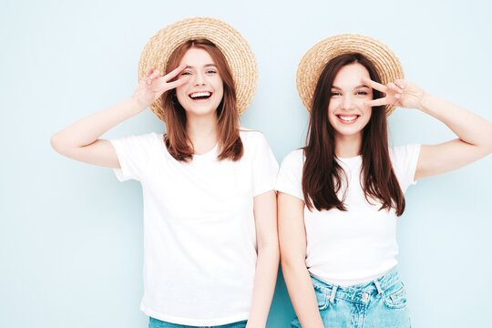 Two young beautiful smiling hipster female in trendy summer white t-shirt and jeans clothes.Sexy carefree women posing near light blue wall in studio.Positive models in hats in sunglasses