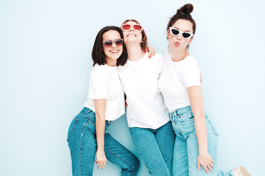 Three young beautiful smiling hipster female in trendy same summer white t-shirt and jeans clothes. Sexy carefree women posing near light blue wall in studio. Cheerful and positive models having fun