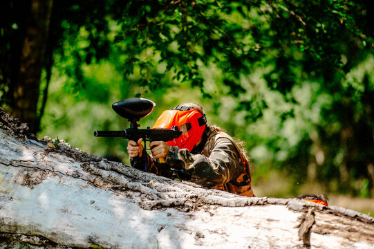 Paintball player in an orange mask hiding behind a fallen tree log and aiming
