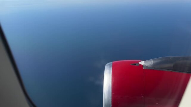 Oblique passenger seat perspective showing red turbofan engine nacelle and partial wing seen through airplane window above open ocean with daylight reflections and faint exhaust vapor