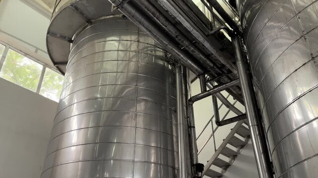 Low upward view between two tall stainless steel storage tanks with dark overhead pipes, metal supports, and central stair structure, showing bright reflected highlights, pale light, and clean heavy