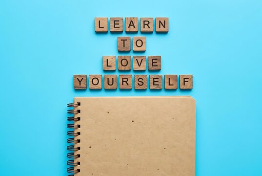 Wooden letters spelling "LEARN TO LOVE YOURSELF" above a notebook on a blue background. Personal growth and self-care concept