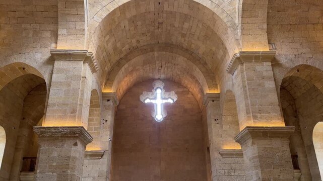 Front-facing view inside historic stone church interior shows glowing cross above altar wall, massive arches, vaulted ceiling, warm uplighting, decorative vessels, and symmetrical sacred space with