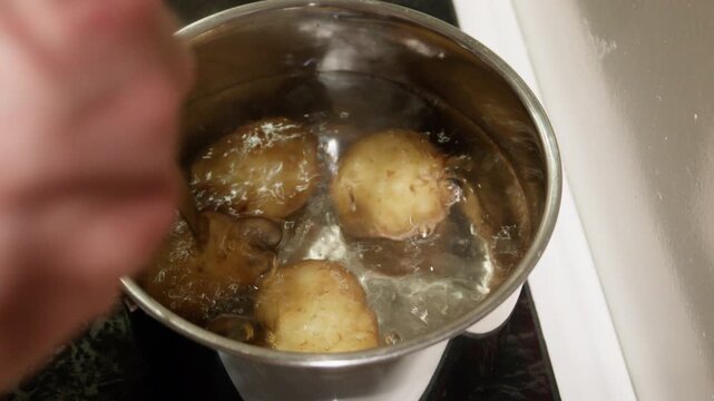 Cook checks three whole potatoes with a fork in a gently bubbling pot on a home stovetop, seen from above.