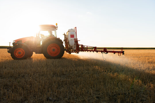 Agricultural tractor spraying harvested field at sunset