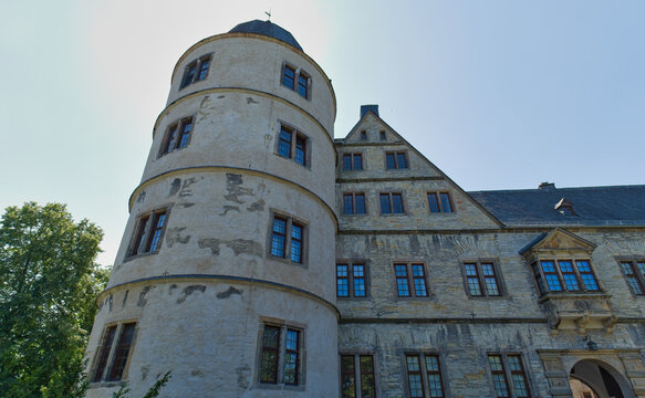 A historic Wewelsburg Castle features a prominent round stone tower with a conical roof, connected to a multi-story building under bright sunlight.