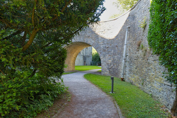 A winding path leads under a historic stone archway, flanked by vibrant green grass and trees. The ancient arch is part of a castle wall - Wewelsburg Castle, Westphalia, Germany © Robert
