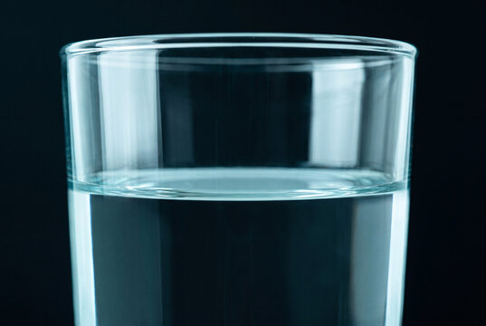A close-up of a clear cylindrical glass filled with water against a dark black background with moody blue lighting.
