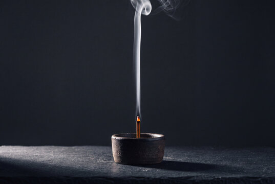A single burning incense stick in a small dark ceramic bowl releases a delicate curl of white smoke against a dark grey background.