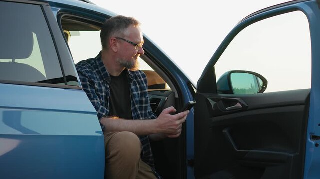 Adult man using a smartphone while sitting in a parked car in the countryside at sunset, capturing remote communication, mobility and a modern lifestyle.