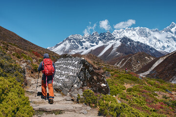 Fototapeta premium Hiker walking on scenic mountain trail past an ancient Buddhist mani stone towards snow-capped peaks in Nepal Himalayas.