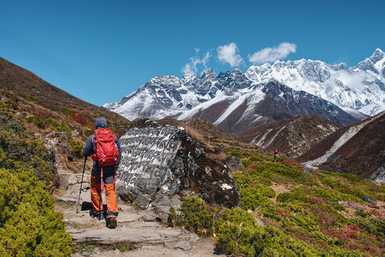 Hiker walking on scenic mountain trail past an ancient Buddhist mani stone towards snow-capped peaks in Nepal Himalayas.