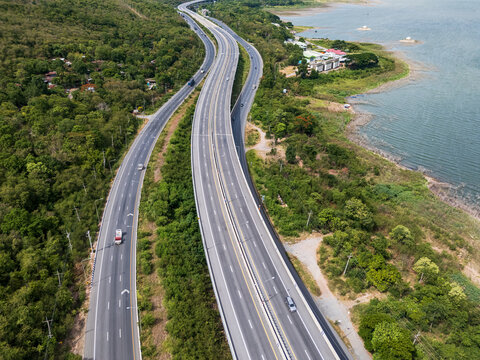 Drone aerial view of a highway running beside a reservoir with lush green mountains, Transportation infrastructure and renewable energy concept
