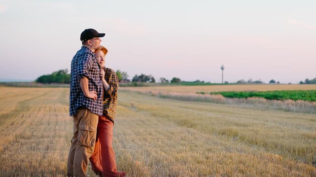 Happy farming couple enjoying a beautiful sunset while standing in a freshly harvested wheat field, celebrating a successful harvest, teamwork and the rewards of rural life.