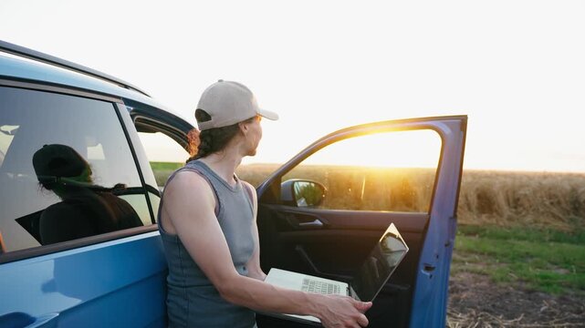 Young professional female farmer using a laptop beside a parked vehicle in the middle of a field, managing farm operations and demonstrating leadership in modern agriculture.