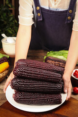 Fresh Black Purple Sticky Corn Cobs on Plate Held by Woman in Kitchen with Vegetables © BuiThi