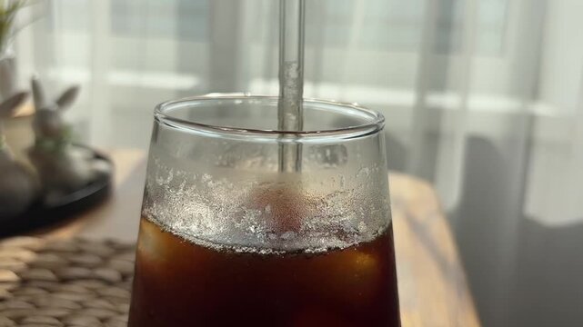 Hand stirring cold iced coffee with a glass straw to espresso in a tall cup, summer drink preparation