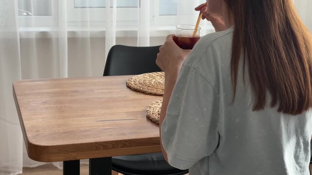 Woman drinking refreshing iced coffee through a straw at home at a table, cozy summer morning relaxation