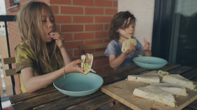 Siblings picking up and eating fresh wraps on a sunny balcony