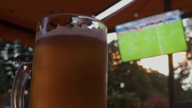 Frosty beer mug before blurred soccer screen in outdoor bar at sunset, warm social atmosphere