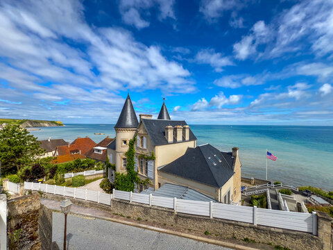Historic coastal villa in Arromanches, Normandy, France, overlooking the sea and D-Day historic coast, with a cyclist on the street and the USA flag waving under a blue sky.