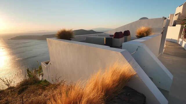 White architecture in Santorini island, Greece. Blue sea and the blue sky at sunset. Beautiful terrace with chaise lounges. Plants swaying in the light wind. Travel and summer vacation concept. 
