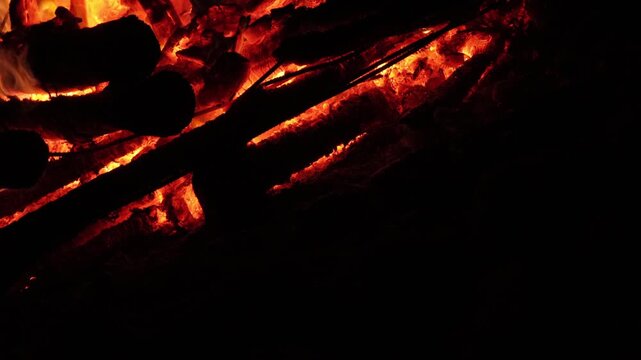 Glowing embers and charred wooden logs in bonfire at night, close-up of burning coals