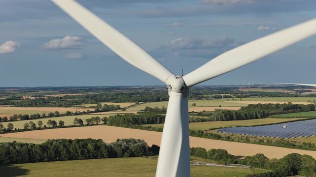 Giant white turbines generate clean electric power above rows of solar panels during a sunny day in rural Germany with fields and forests visible under a blue sky with soft white clouds.