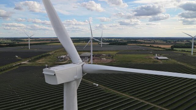 Large white wind turbines rotate over an extensive field covered with rows of solar panels during a sunny afternoon to provide renewable energy for the local power grid in the countryside