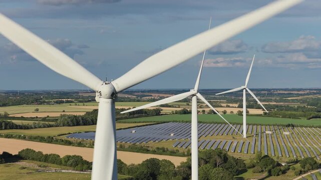 White wind turbines rotate over rows of solar panels in a green field under a clear blue sky during the bright afternoon in England to generate clean renewable electricity for the local grid