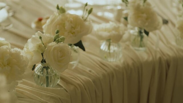 Medium shot of a row of small glass vases filled with fresh white roses and peonies arranged along a banquet table