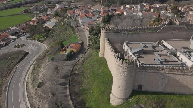 A drone shot on a winter day, panning right over Maqueda Castle&mdash;featuring its medieval walls, battlements, and an inner courtyard with modern facilities&mdash;with the village of Maqueda visible