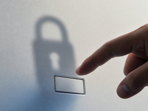 Close-up of a hand approaching a blank physical button while a padlock-shaped shadow appears behind it, representing secure login, authentication and access control.