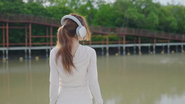 Active female athlete putting on headphones, runner getting ready for outdoor jog near water, wearing white sportswear with wooden bridge, green forest in background, enjoying music.