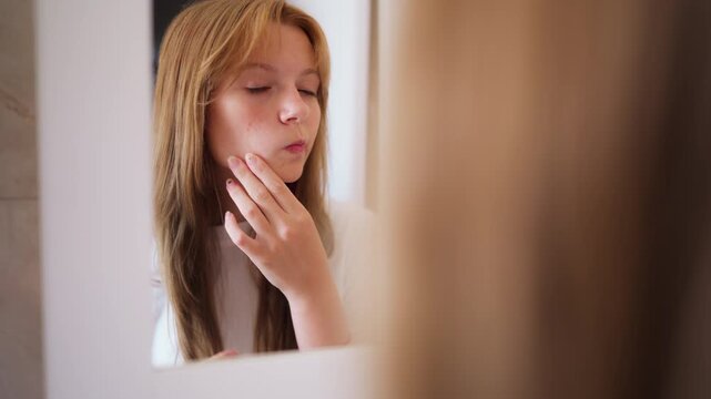 Teenage girl examining acne-prone skin and squeezing pimples while look in bathroom mirror during daily skincare routine. Close-up teenage acne, puberty, facial blemishes, skin concerns, dermatology