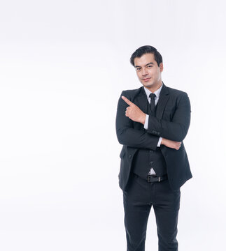 Confident handsome businessman in dark suit standing on white background pointing finger aside at copy space. Professional executive presenting corporate promotion or advertising concept.