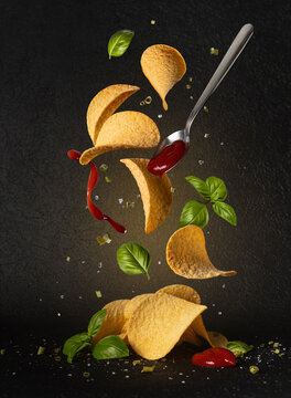 Potato chips falling with ketchup, basil leaves and spices. Food levitation concept on black background