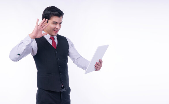 Happy handsome businessman in dark vest waving hand to tablet screen making video call on white background with copy space. Professional executive in online meeting or virtual conference.